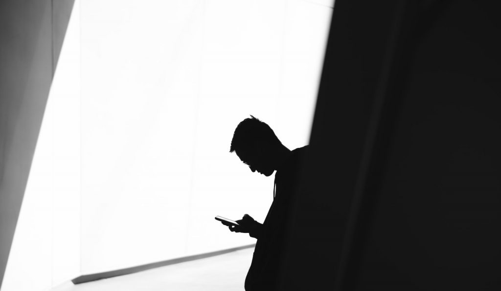 Image of a sillhouette of a man on his phone, against a sheer white background. He looks like he's up to something very secretive!